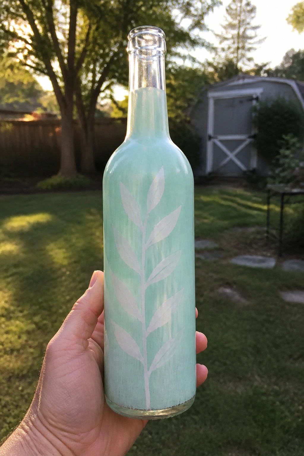 Hand holding mint green bottle with white leaf design in backyard