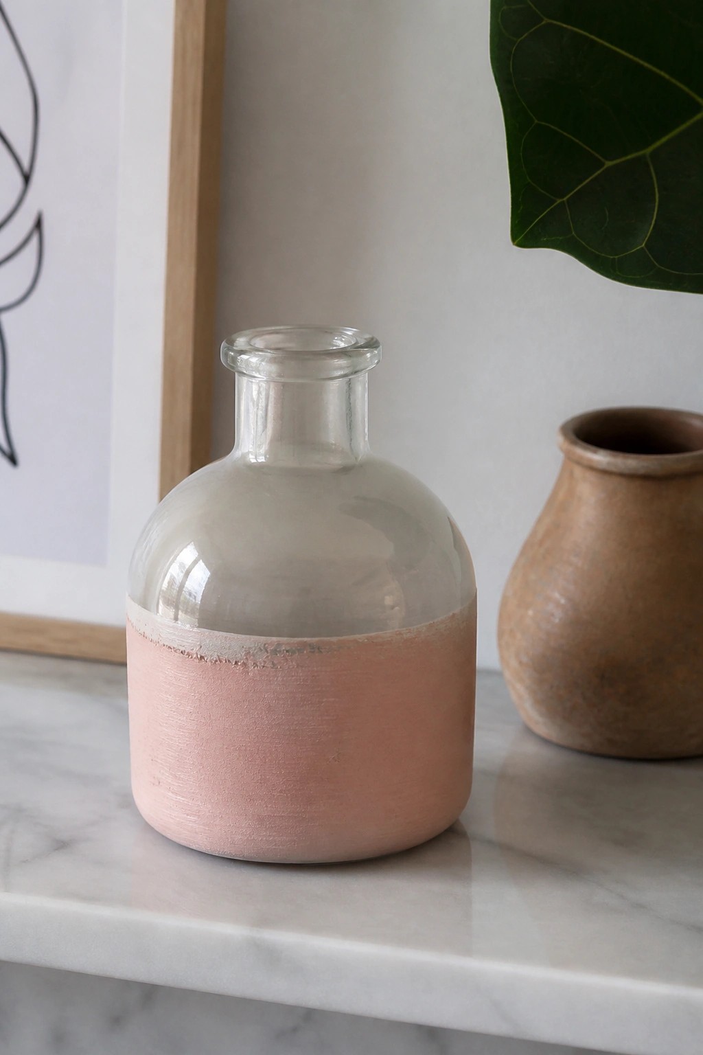 Pink-painted glass bottle beside terracotta pot on white marble surface