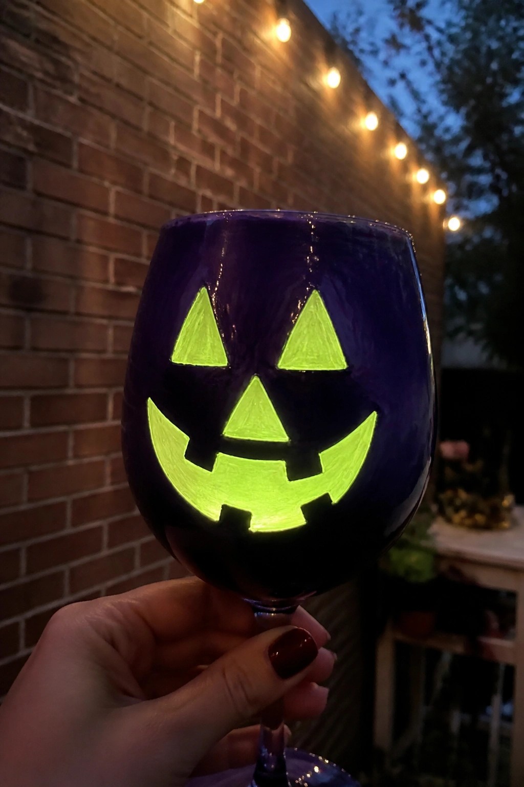 Hand holding purple wine glass with glowing green jack-o'-lantern face, brick wall and lights behind