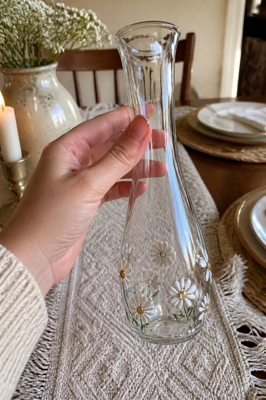 Hand in beige sweater holding clear glass vase with white daisy decorations