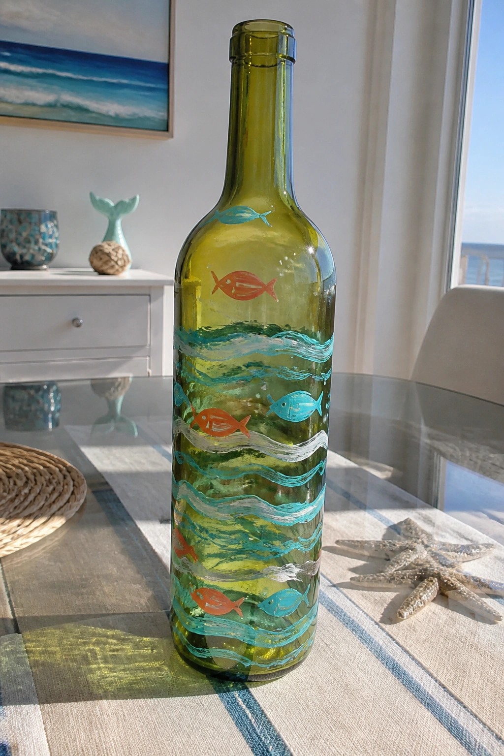 Green glass bottle painted with fish and wavy lines beside starfish on table
