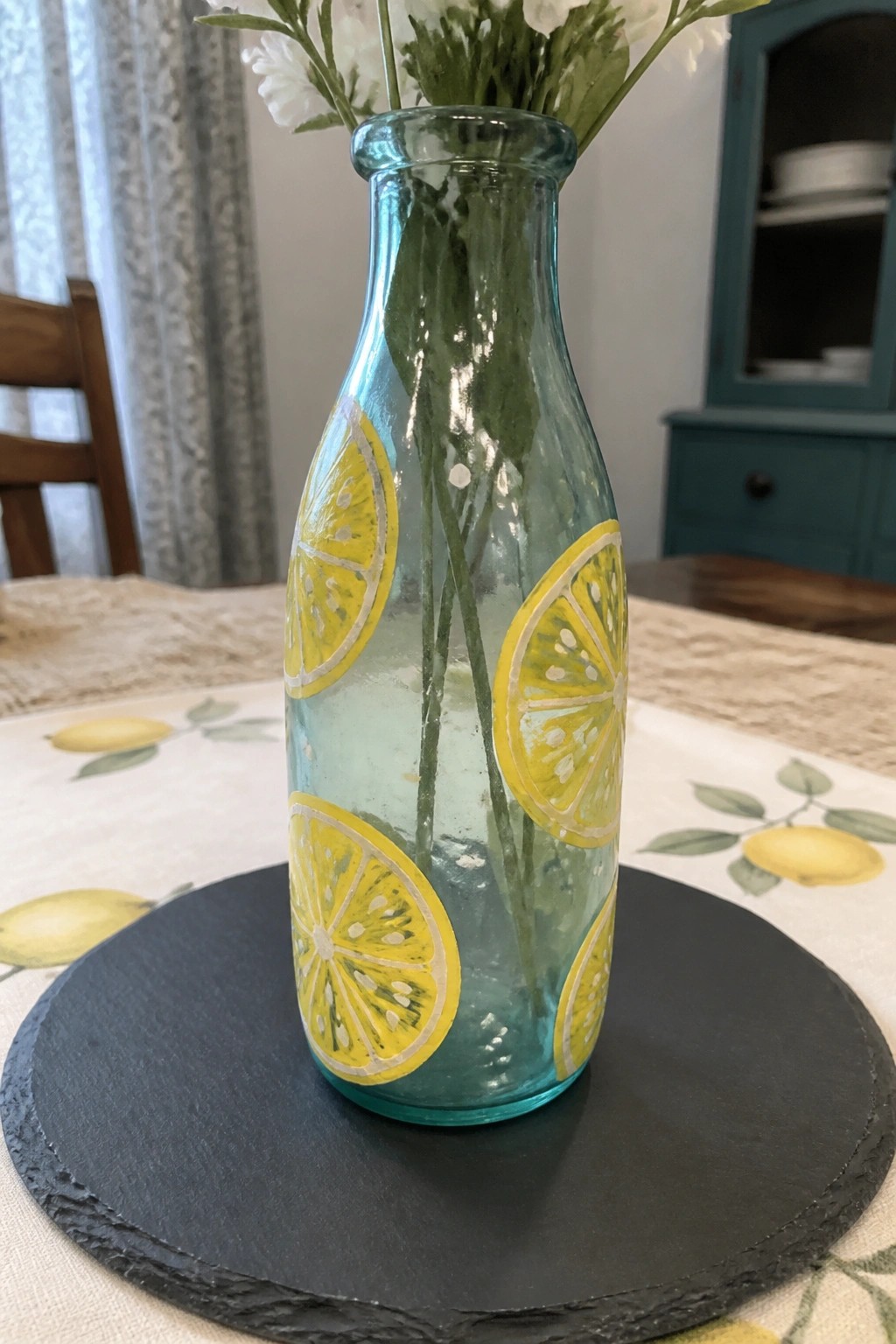 Turquoise glass bottle painted with yellow lemon slices holding white flowers on black tray