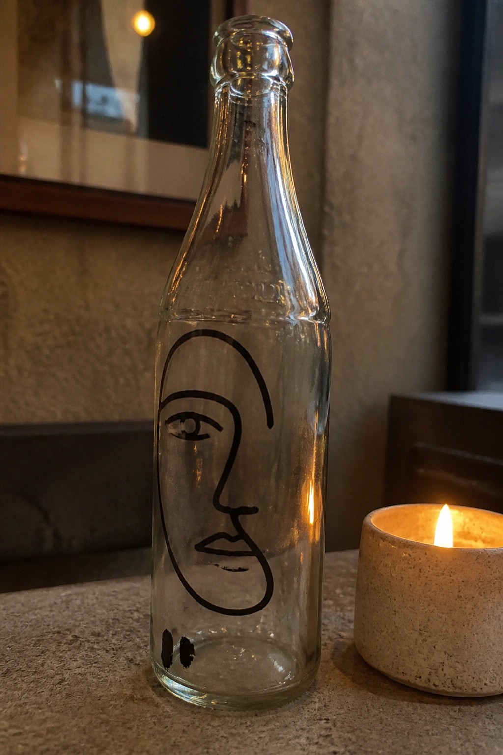 Clear glass bottle with line-drawn face beside lit candle in holder