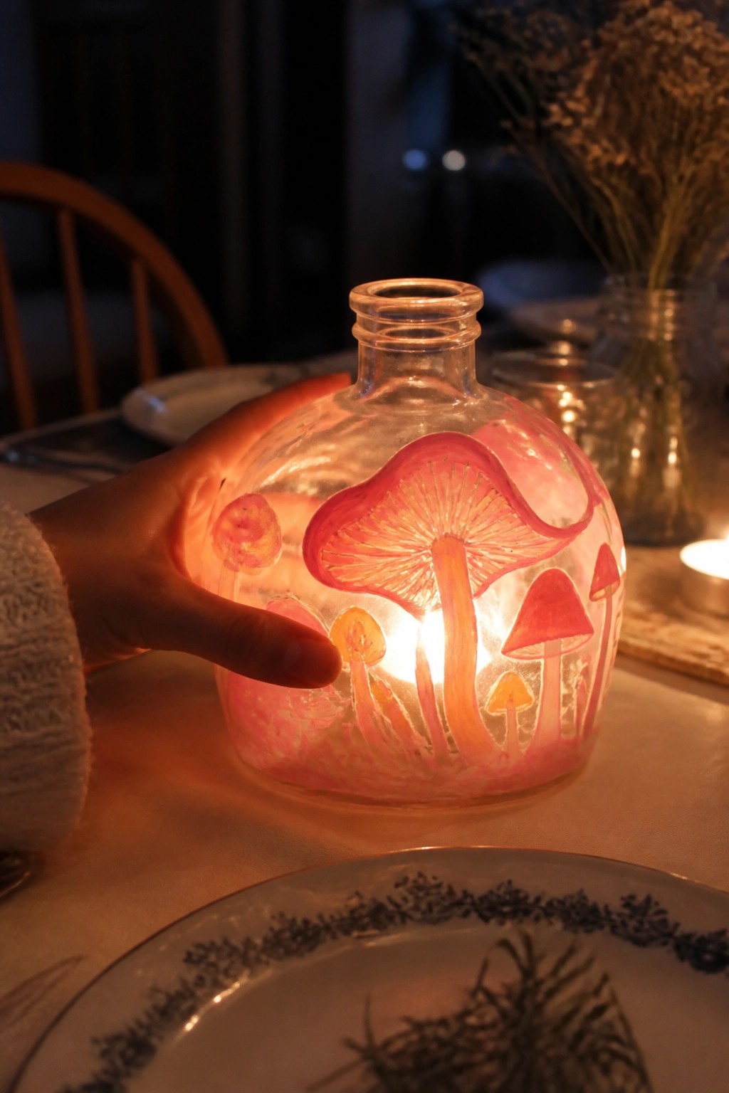 Hand in white sweater holding glowing glass bottle painted with pink mushrooms on table