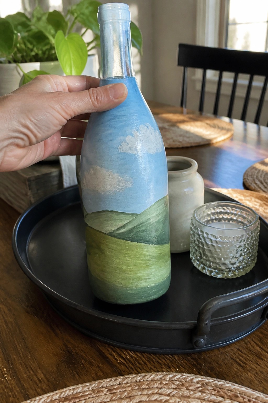 A hand holds a glass bottle painted with blue sky, white clouds, and green hills on a black tray.