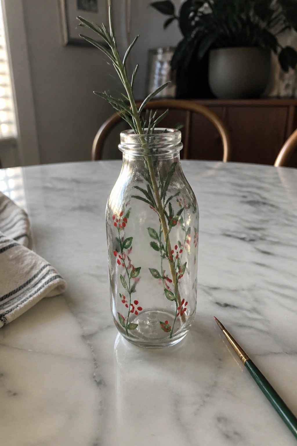 A clear glass bottle painted with green leaves and red berries sits on a marble table with a sprig of rosemary inside and a paintbrush nearby.