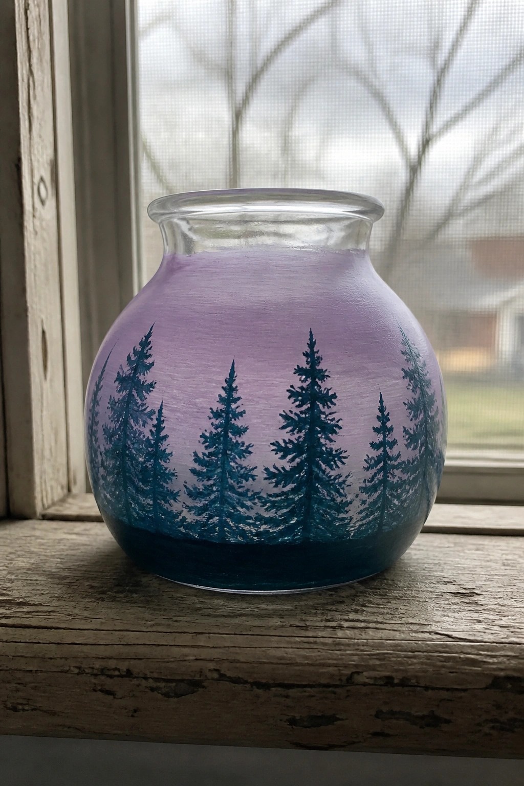 Glass jar painted purple to blue with dark pine tree silhouettes on windowsill.