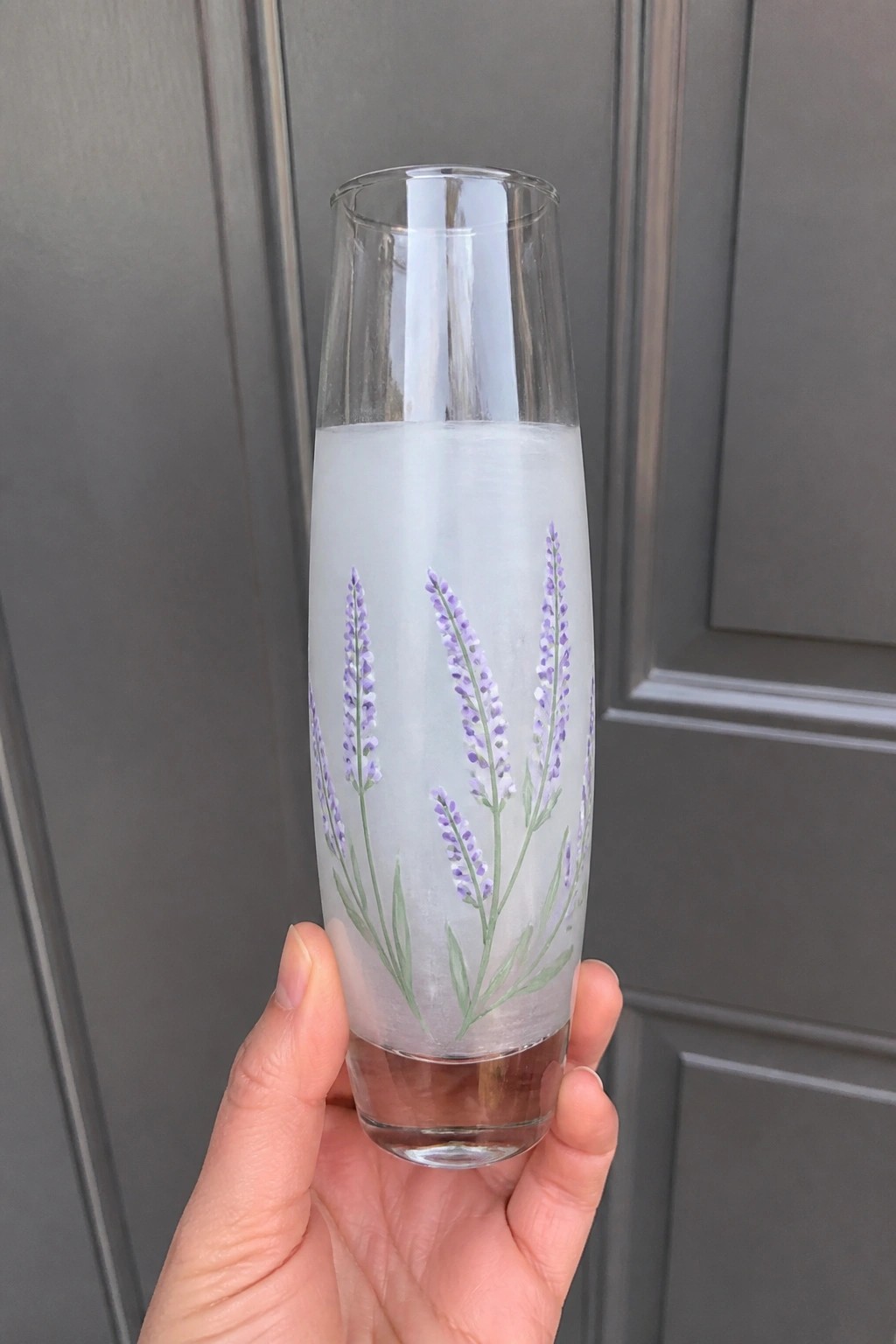 Hand holding tall glass painted with purple lavender flowers and white liquid.