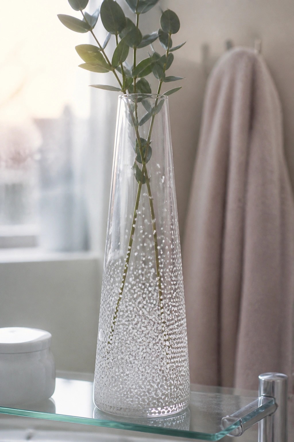 Tall textured glass vase with eucalyptus on shelf beside pink towel