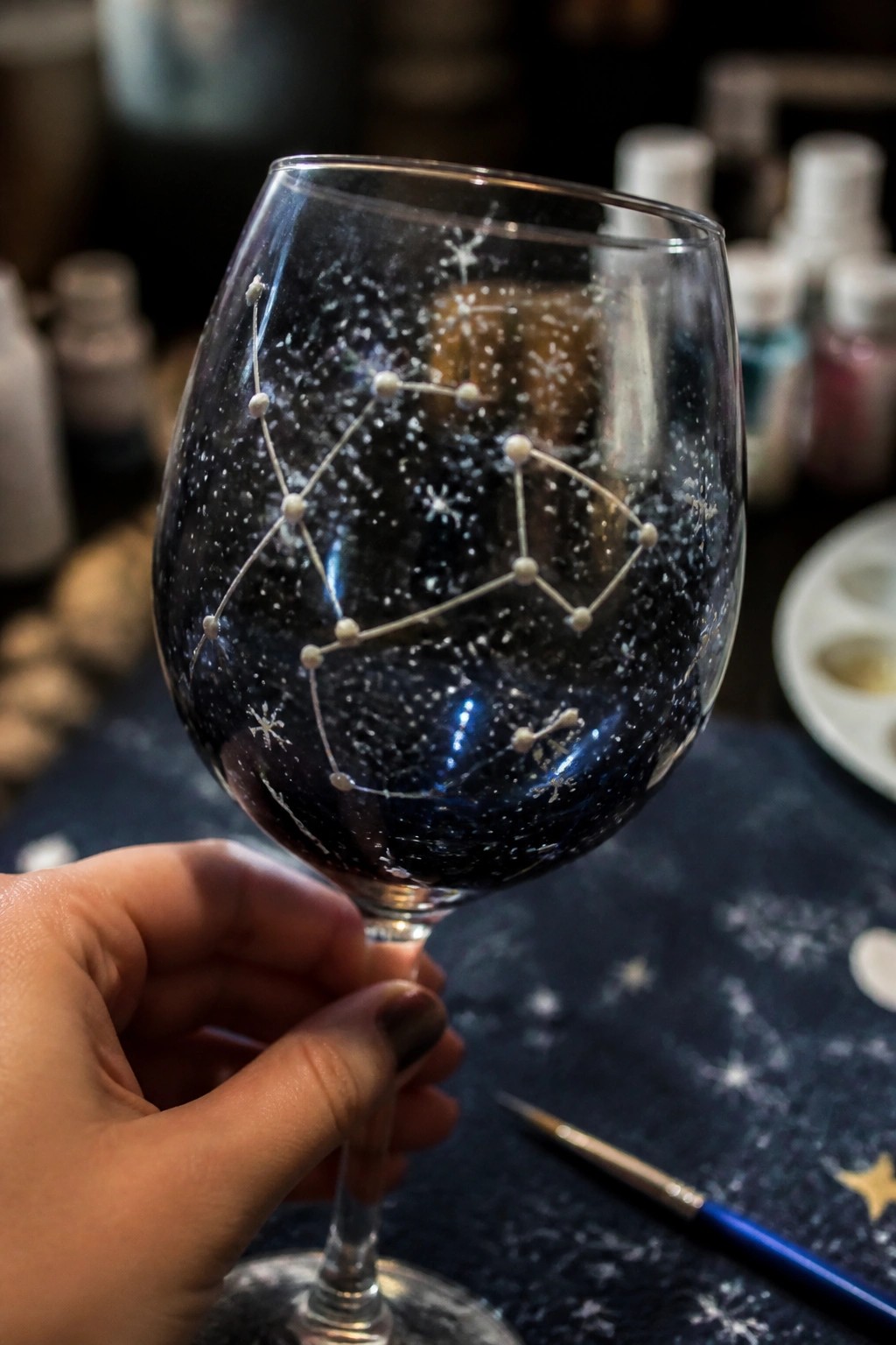 Hand holding wine glass painted with white constellation on dark starry background
