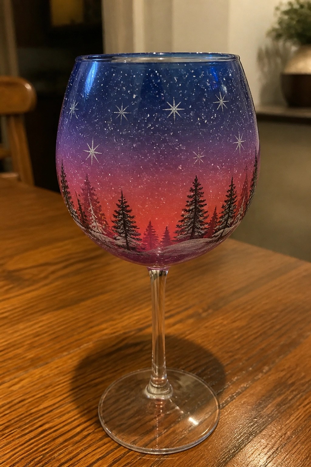 Painted wine glass with starry night sky and snowy pine trees on wooden table