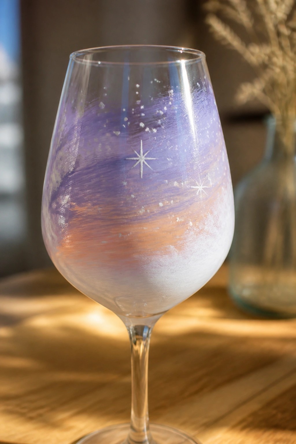 Painted wine glass with purple-orange swirls, white speckles and star designs on wood.