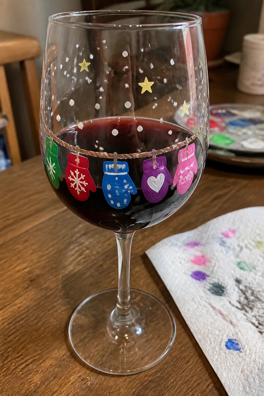 Wine glass of red wine painted with colorful hanging mittens, stars, and dots.