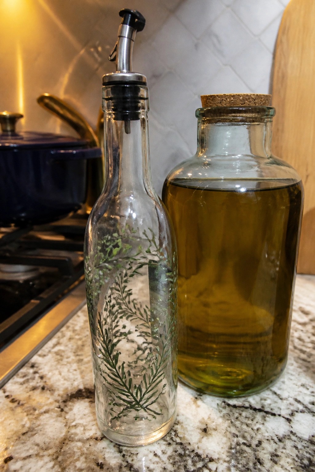 Two glass bottles on marble counter: one with green plant design and pourer, one with oil and cork.
