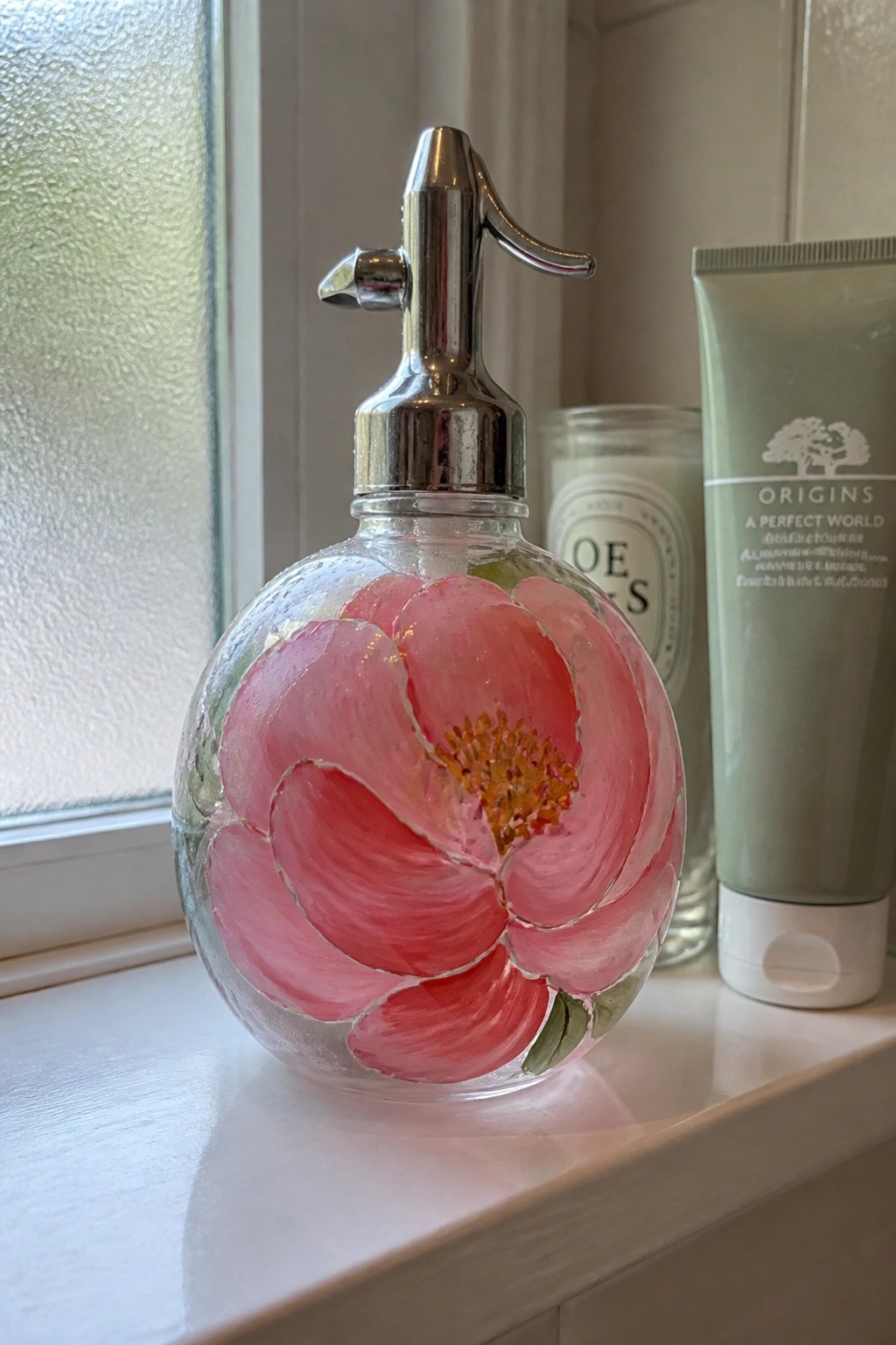 Glass seltzer bottle with pink flower painting beside green Origins tube on windowsill