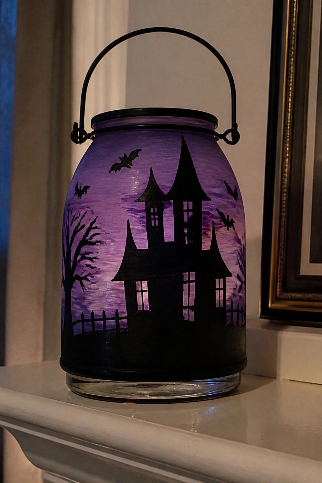 Lit purple glass jar with black haunted house, bats, trees, and fence silhouette