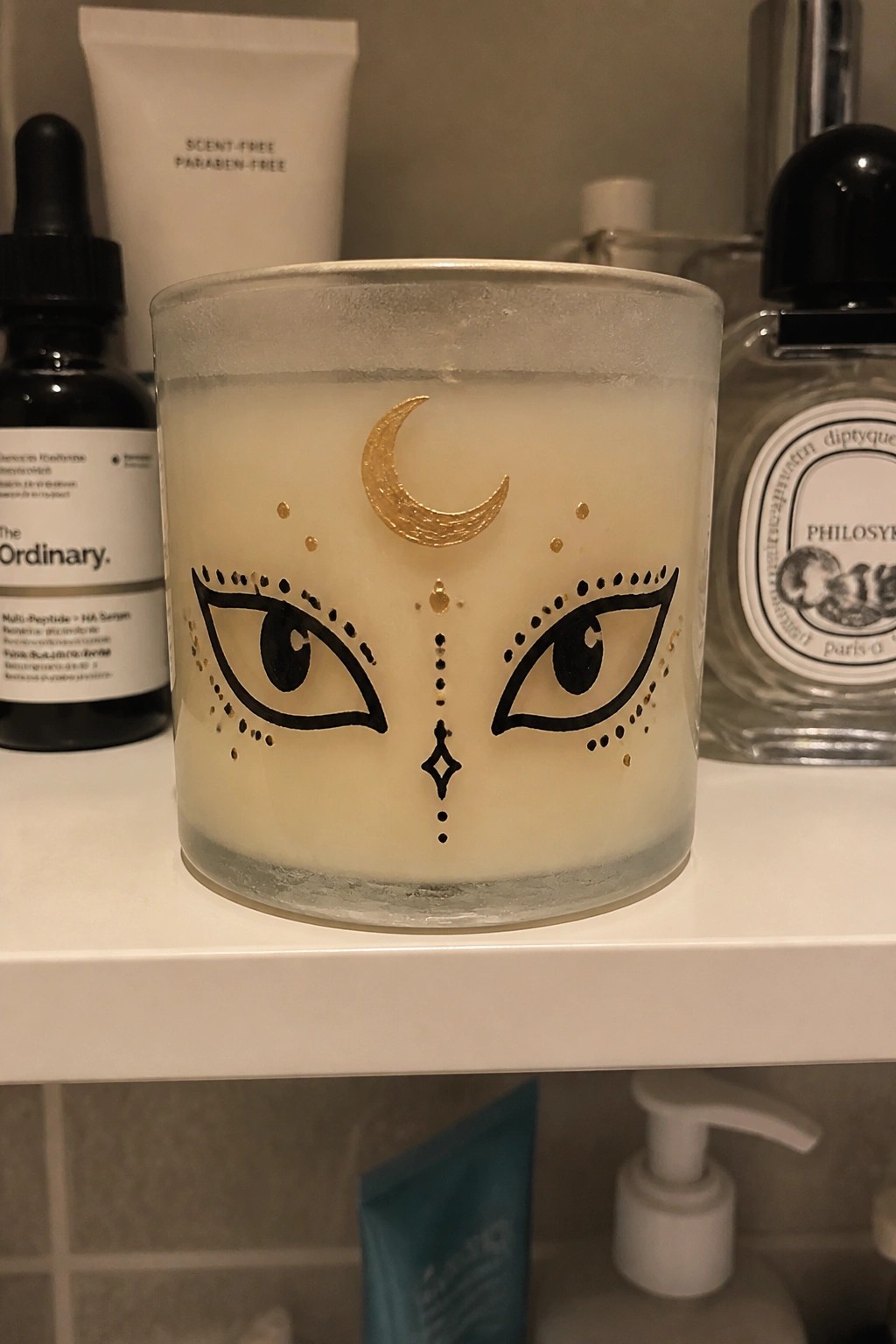 Candle with golden crescent moon, black eyes, and dots on shelf with cosmetics