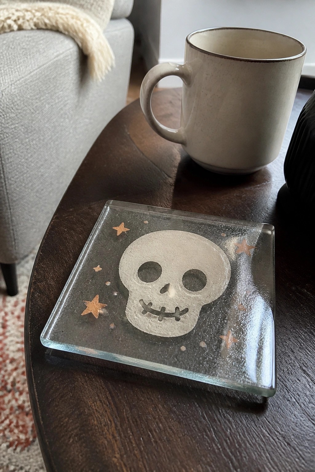 A square clear glass coaster with a white skull and scattered gold stars rests on a dark wooden table beside a beige mug.