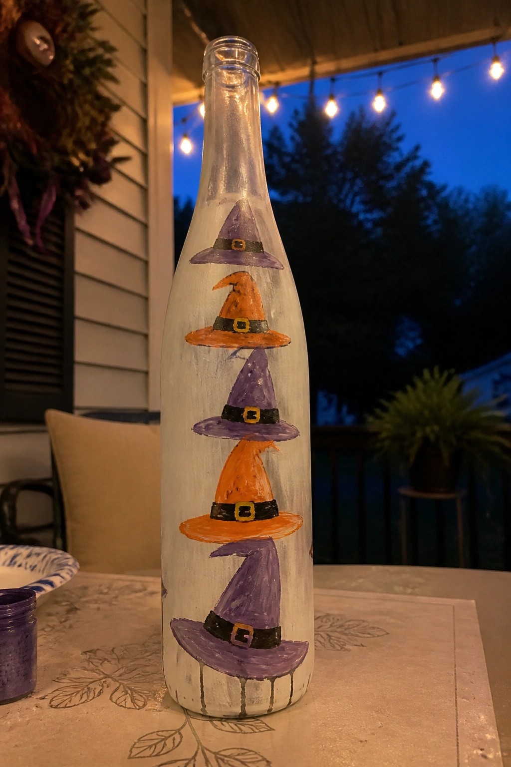 Painted white bottle with stacked purple and orange witch hats on patio table.