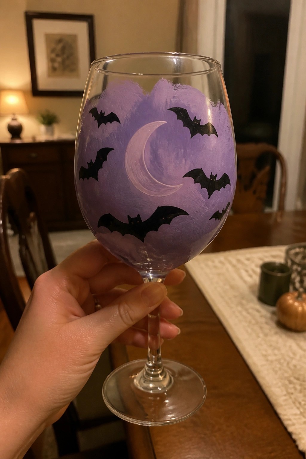 Hand holding wine glass painted purple with white moon and black bats.