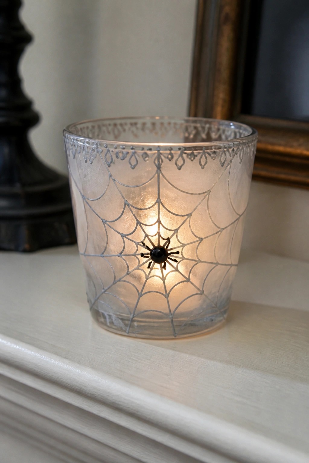 Glowing glass votive etched with spiderweb holds black spider on white mantel