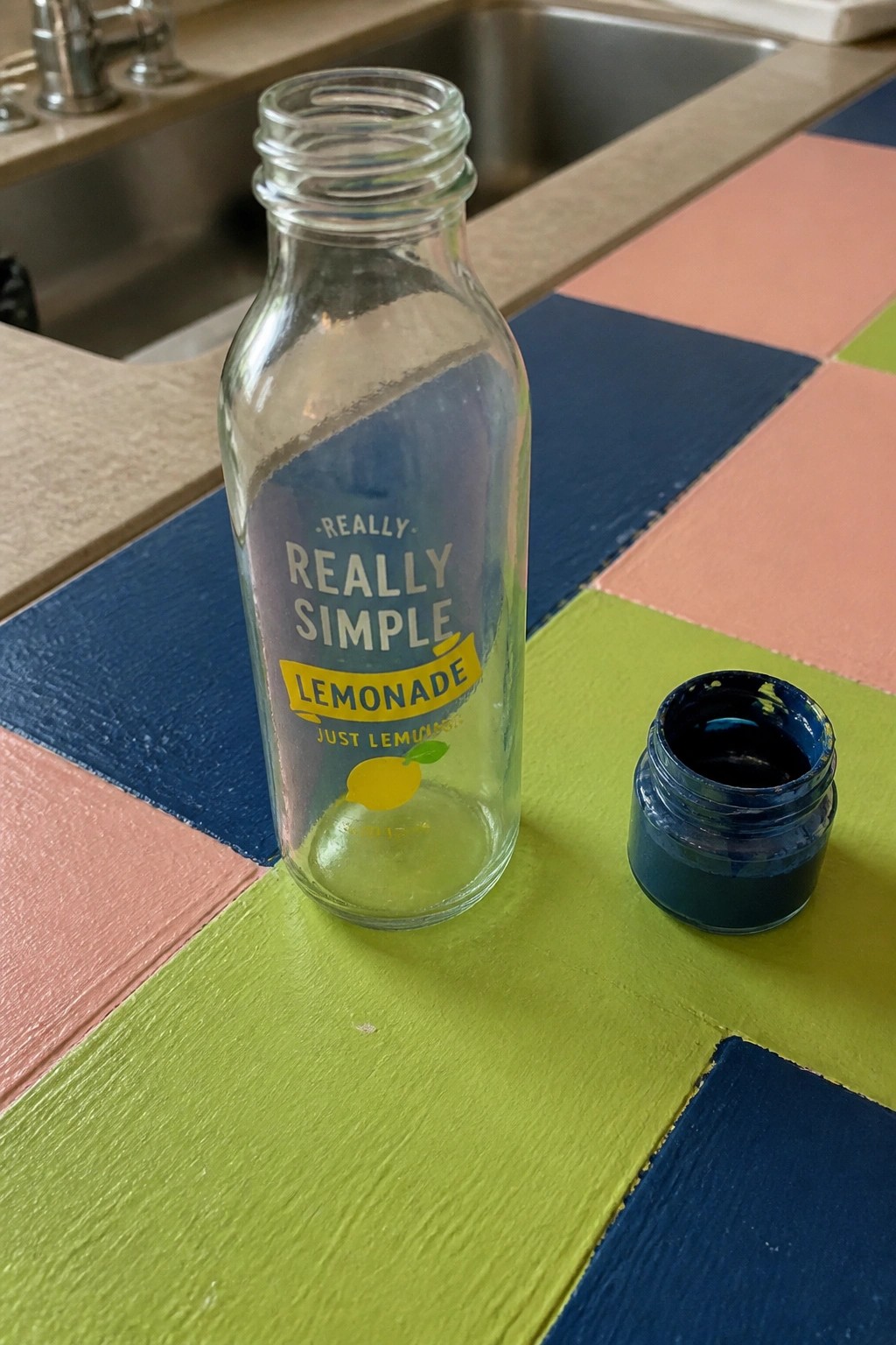 Clear glass lemonade bottle beside open jar of blue paint on colorful wooden surface.