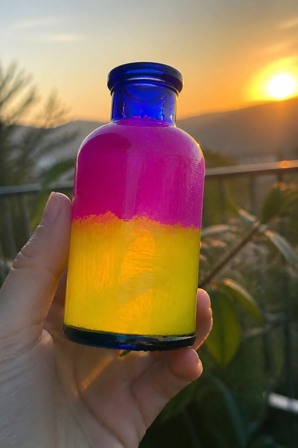 Hand holding pink-and-yellow painted blue glass bottle at sunset