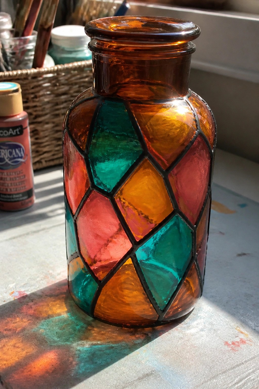 Amber glass vase with colorful mosaic panels on sunlit paint-splattered table.