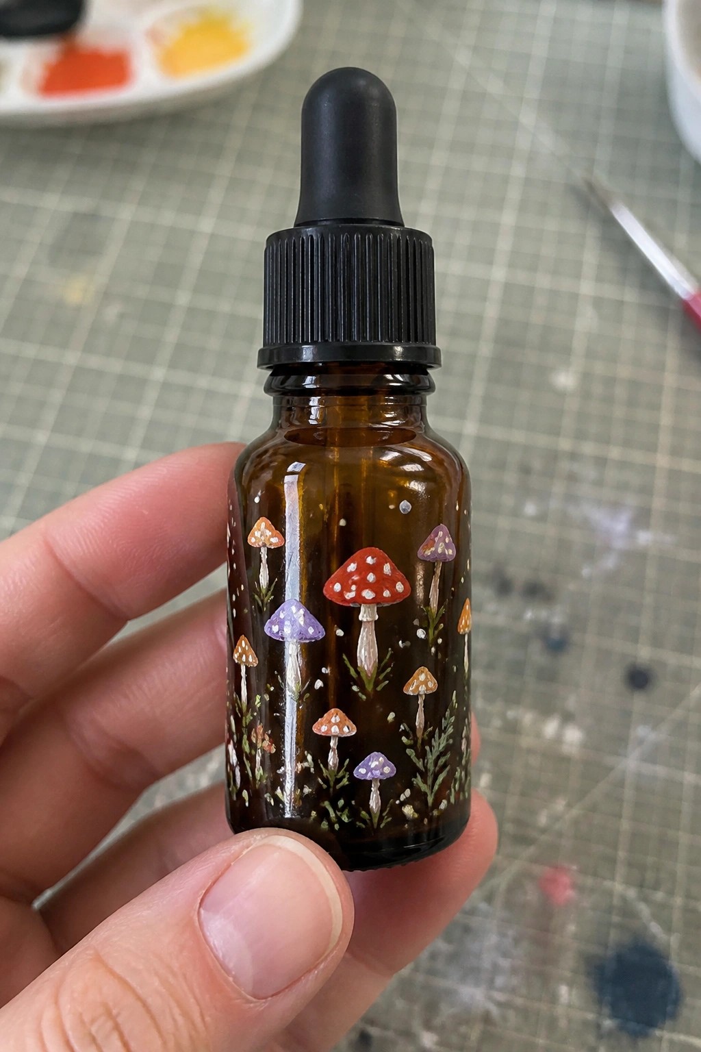 Hand holding amber dropper bottle painted with red, purple, and orange mushrooms.