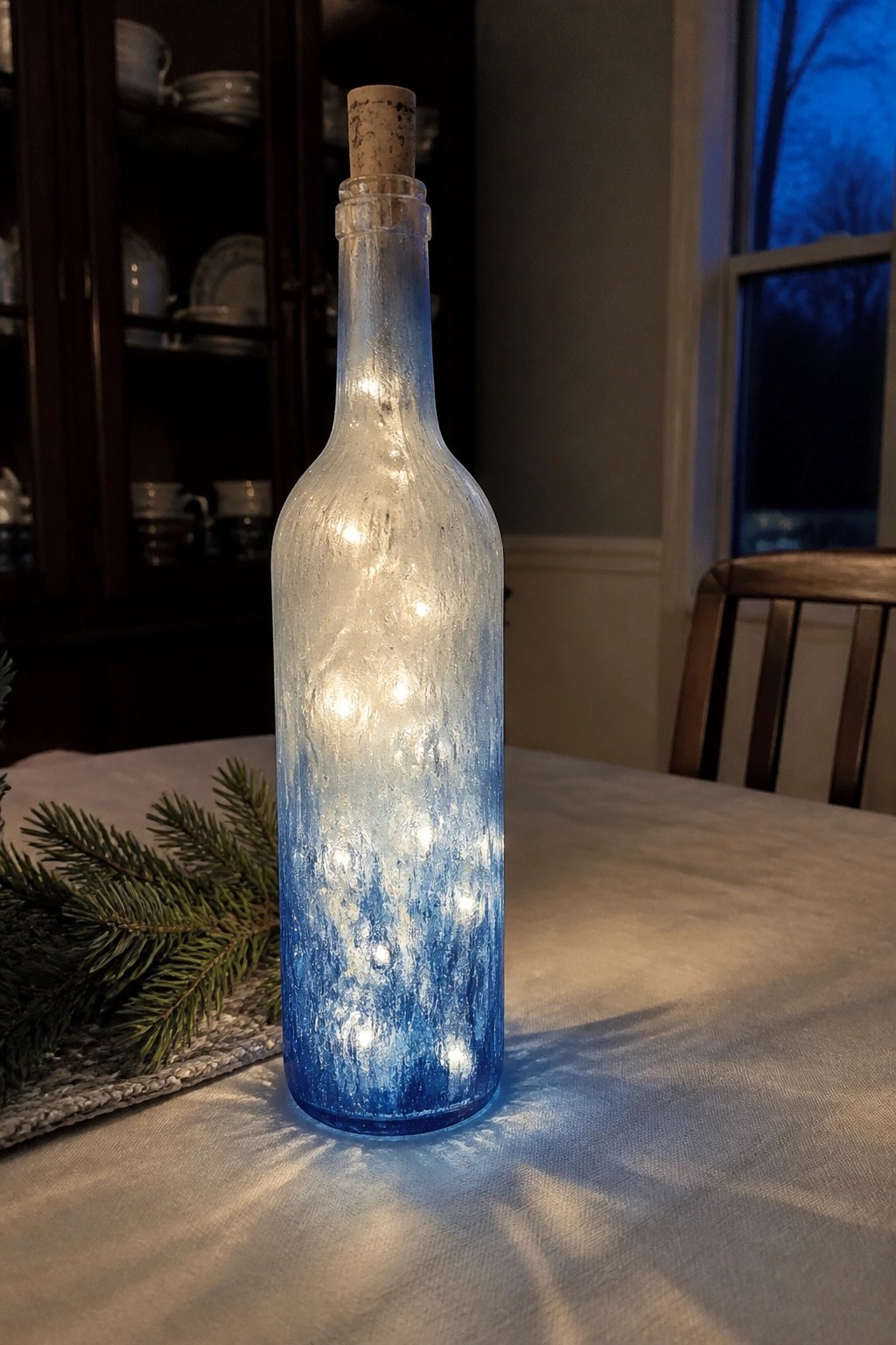 Glowing blue gradient bottle with fairy lights and cork on table beside pine branch