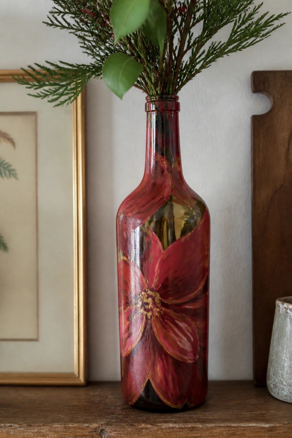 Red painted bottle vase with floral design holding green foliage on wooden table
