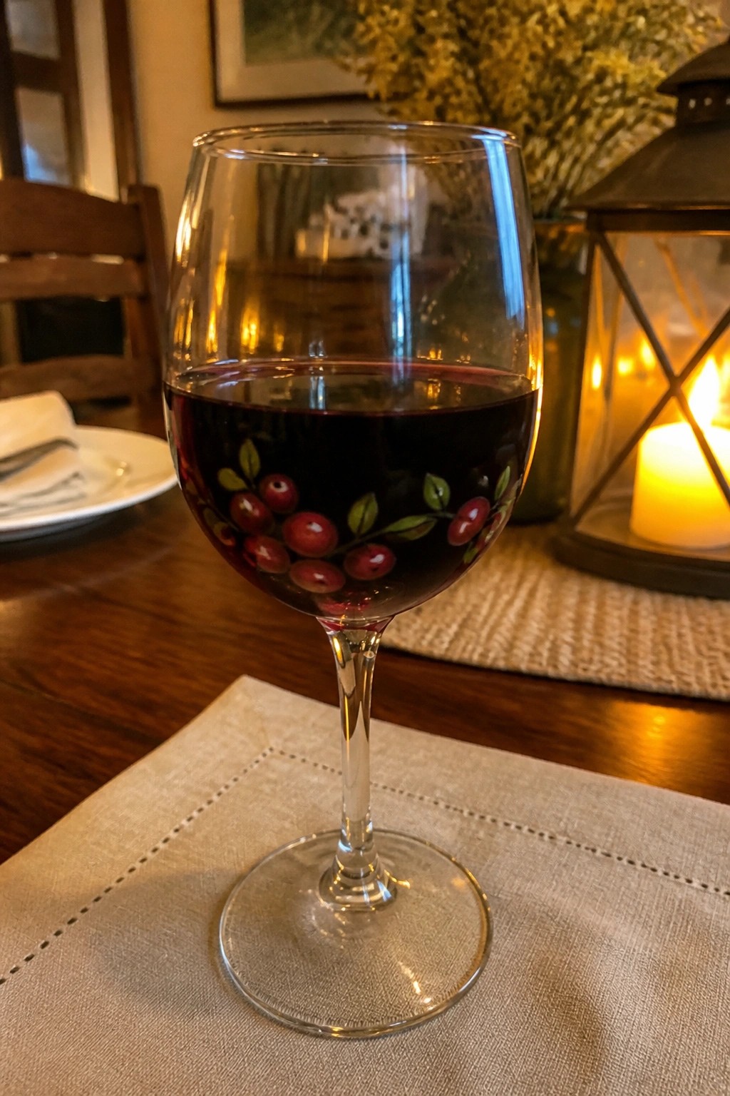 Red wine glass painted with cranberries beside glowing lantern on wooden table