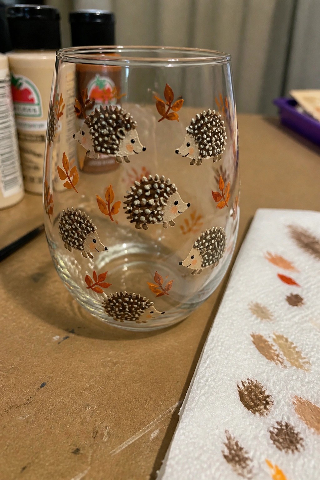 Stemless glass painted with hedgehogs and orange leaves amid paint supplies on wood.