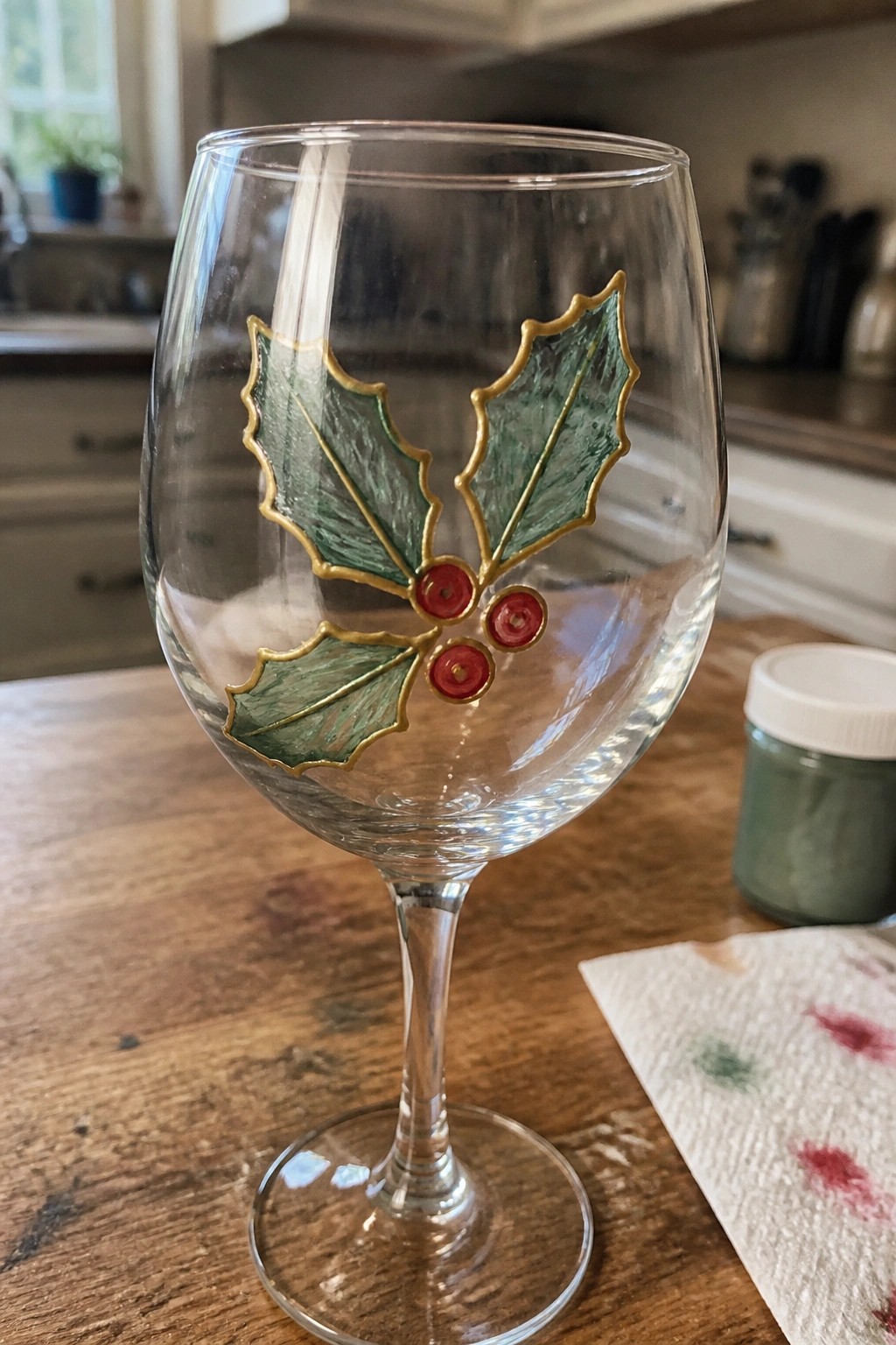 Wine glass with green holly leaves, gold outlines, and red berries on wooden table.