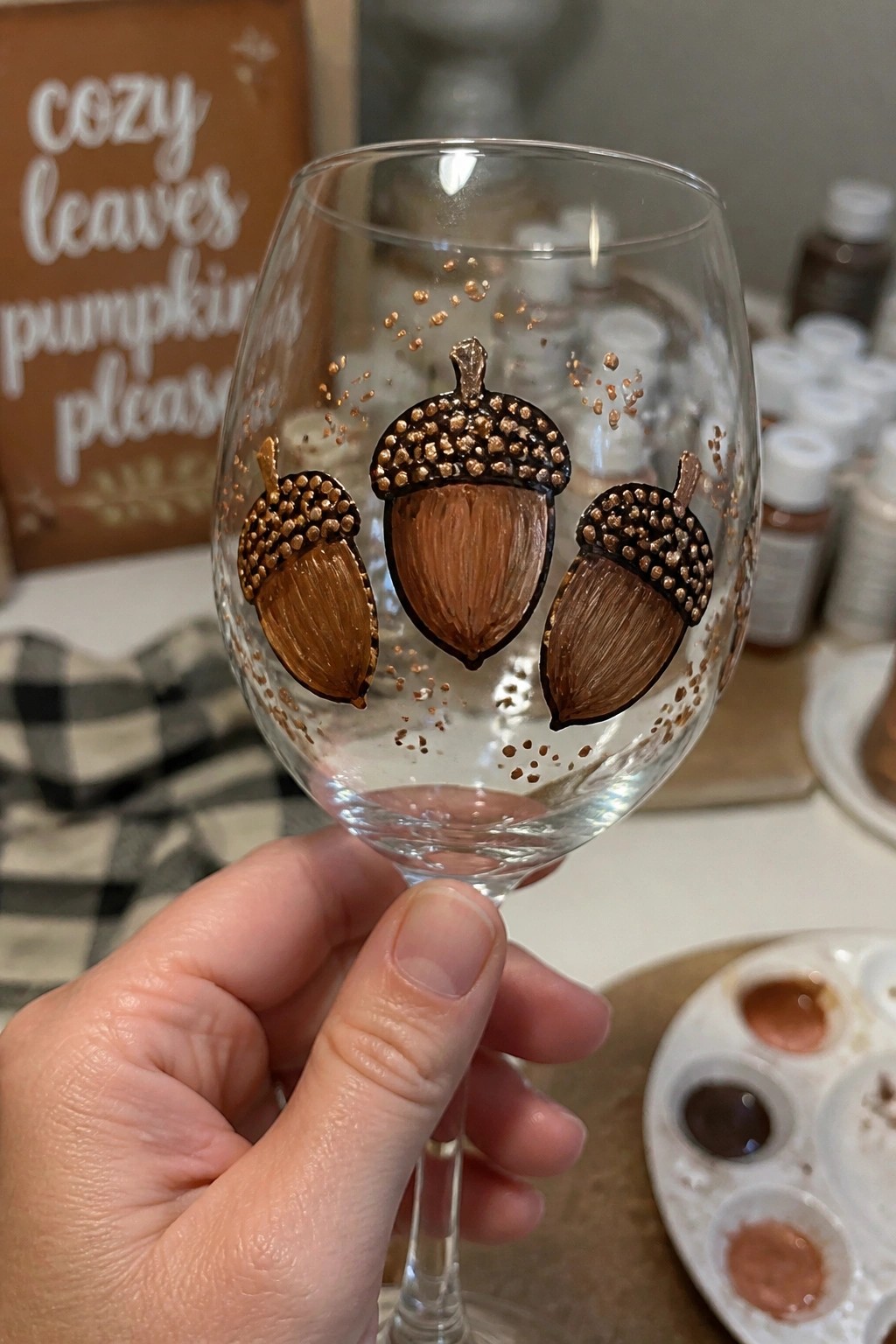 Hand holding wine glass painted with three brown acorns and copper glitter dots