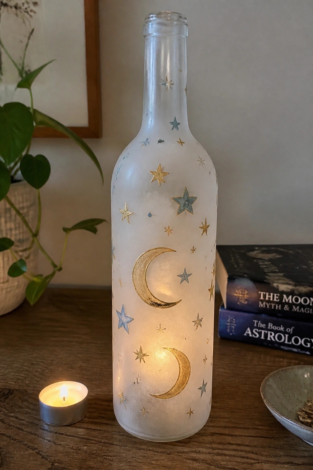 Illuminated frosted bottle with gold moons and stars beside a lit candle on wood.