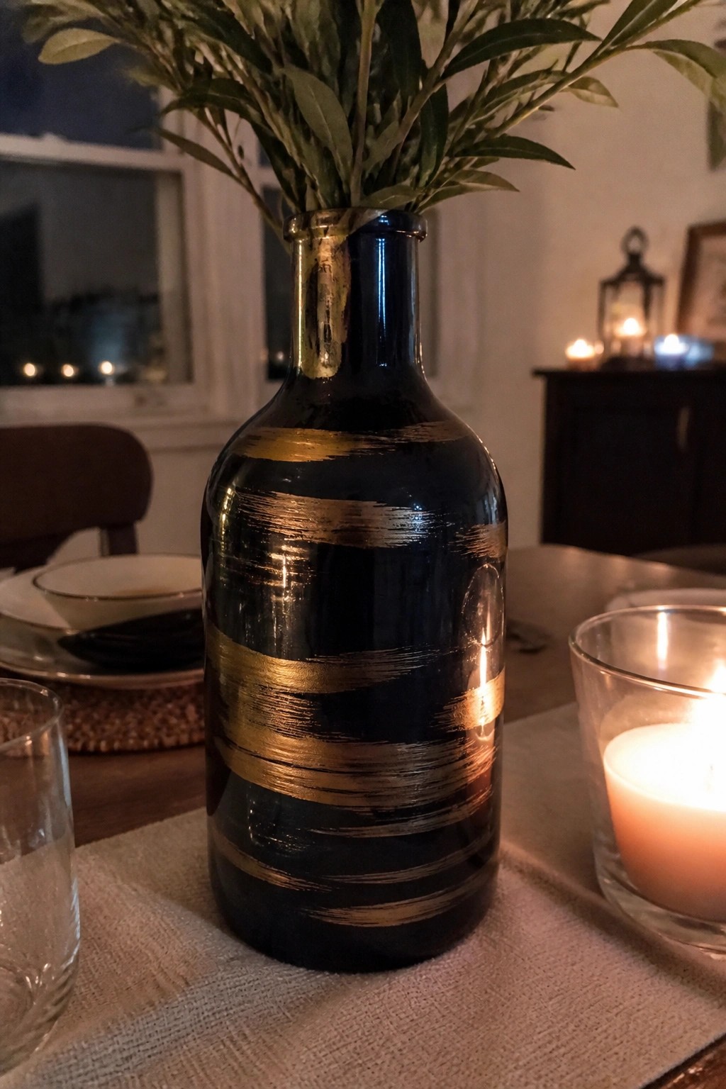Dark blue vase with gold stripes holding greenery beside lit candle on table