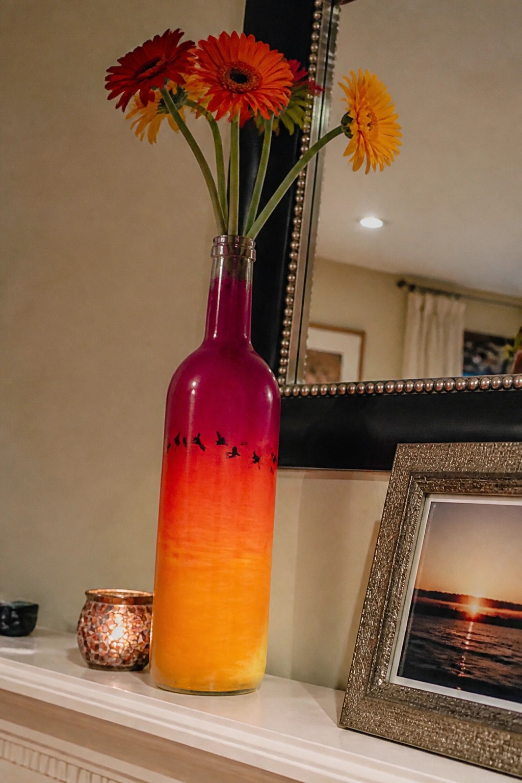 Ombre magenta-orange bottle with bird silhouettes holding red and yellow gerbera daisies on mantel