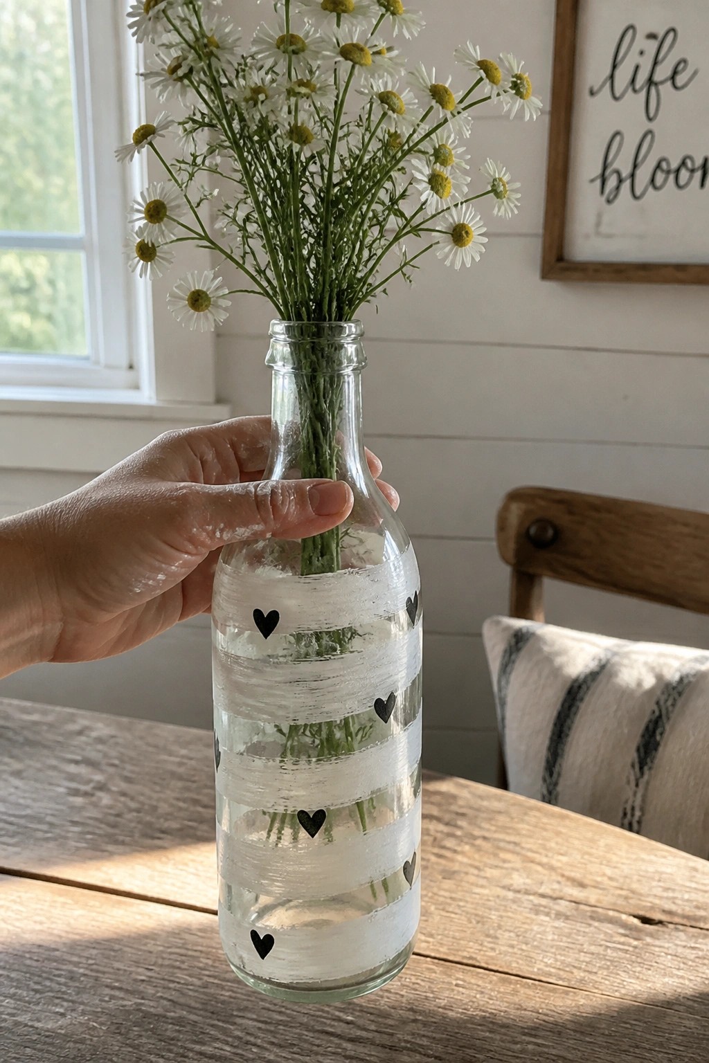 Hand holding glass bottle with white stripes and black hearts filled with daisies on table