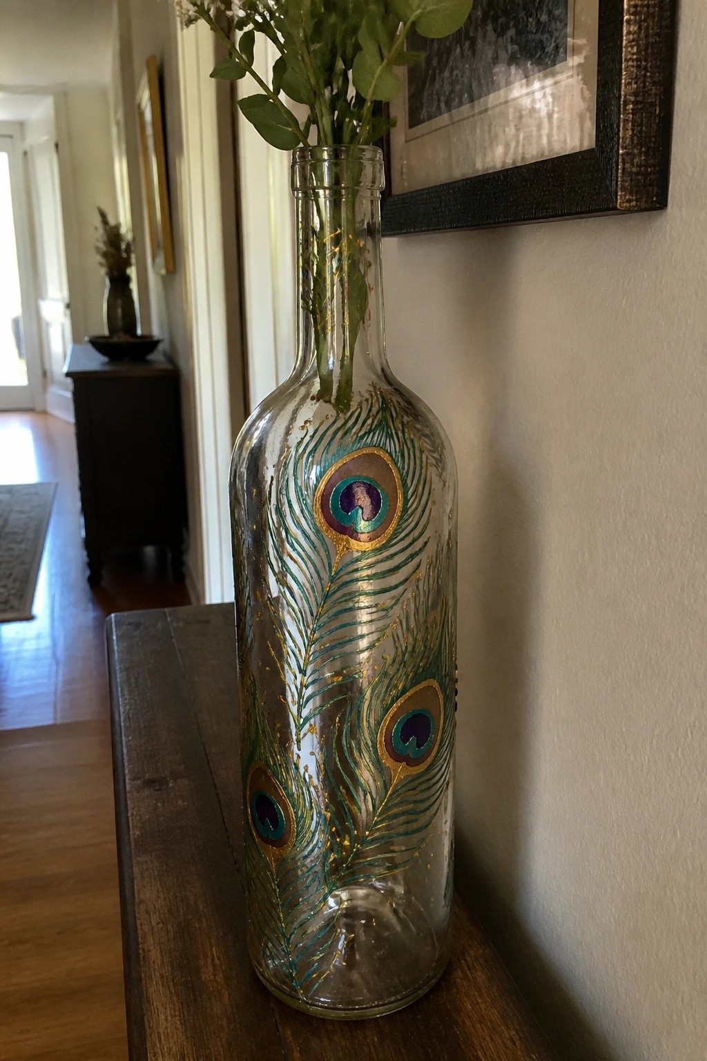 Painted glass bottle with peacock feathers in teal, purple, and gold holding greenery.