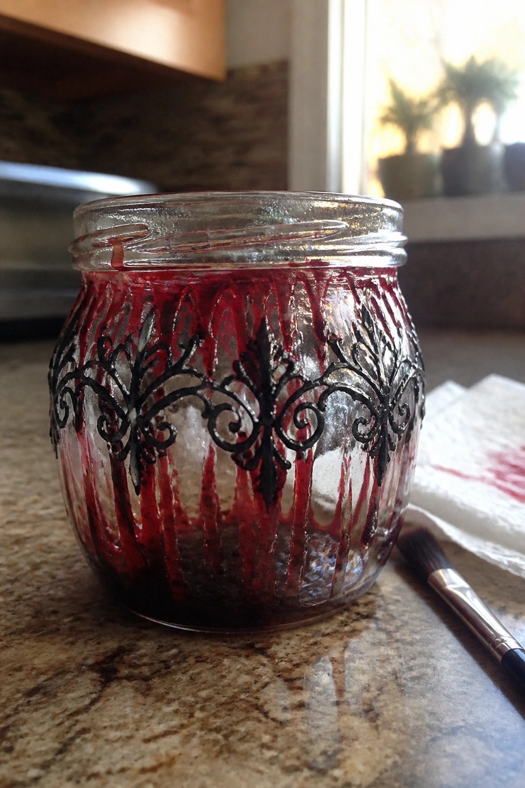 Ornate black-patterned glass jar with red drips beside paintbrush on counter