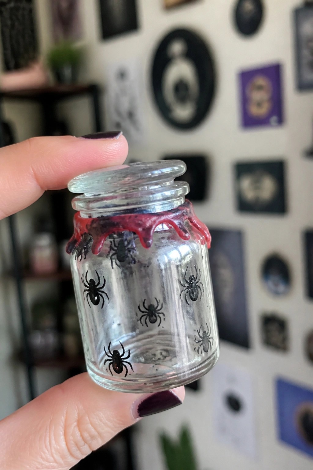 Hand with dark purple nails holds glass jar with black spiders and red drips.