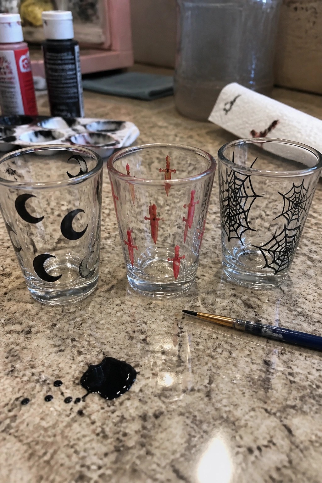 Three hand-painted glasses with moons, swords, and spiderwebs amid paint supplies on countertop.