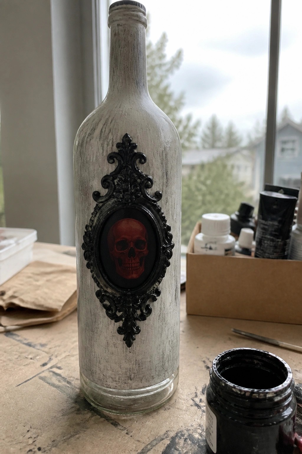 A white painted glass bottle with a black ornate frame containing a red skull, sitting on a wooden table with art supplies nearby.