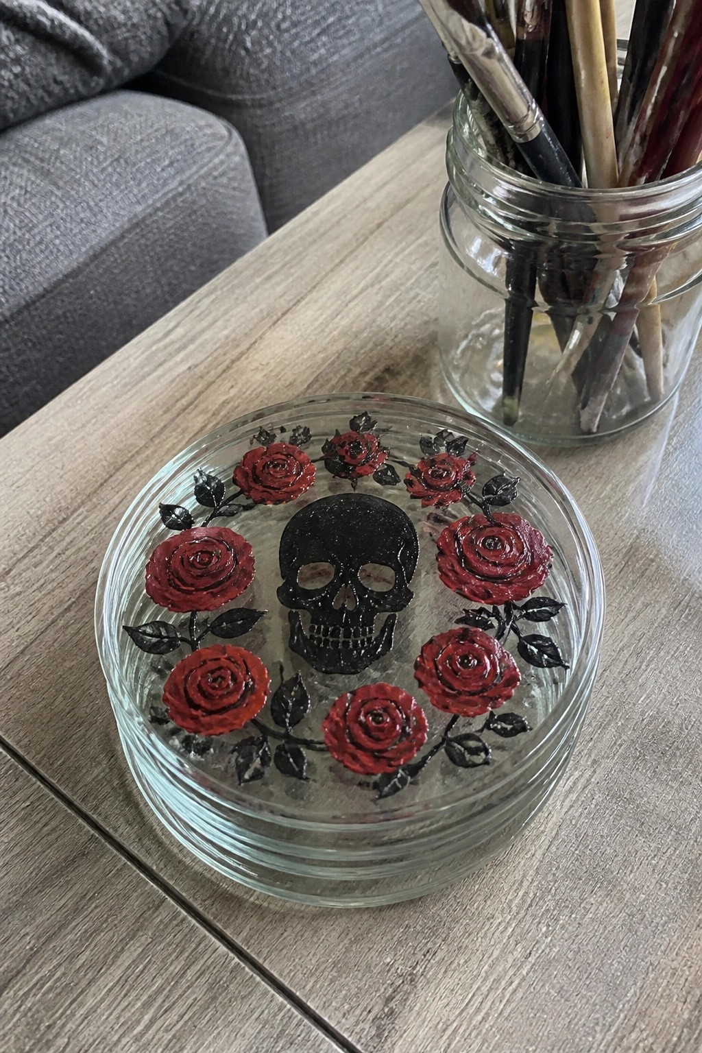 Glass coaster with black skull centered amid red roses and black leaves on wood