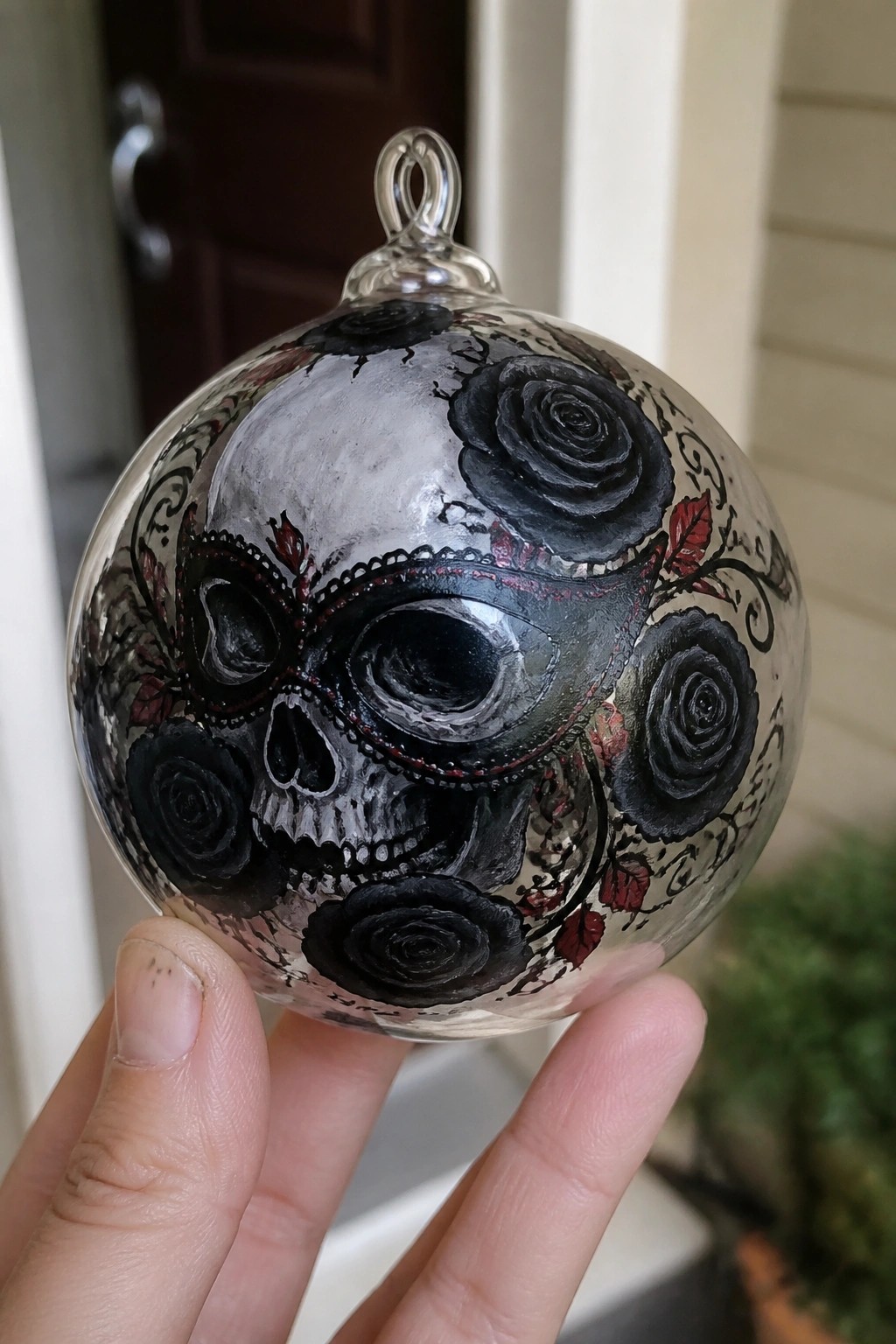 A hand holds a clear glass spherical ornament painted with a skull wearing a black masquerade mask surrounded by black roses and red leaves.