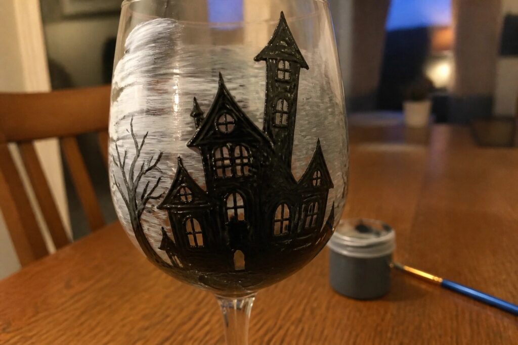 24 Haunted House Wine Glass Painting Ideas That Create a Spooky Centerpiece Wine glass hand-painted with black haunted house, windows, trees, and fence.