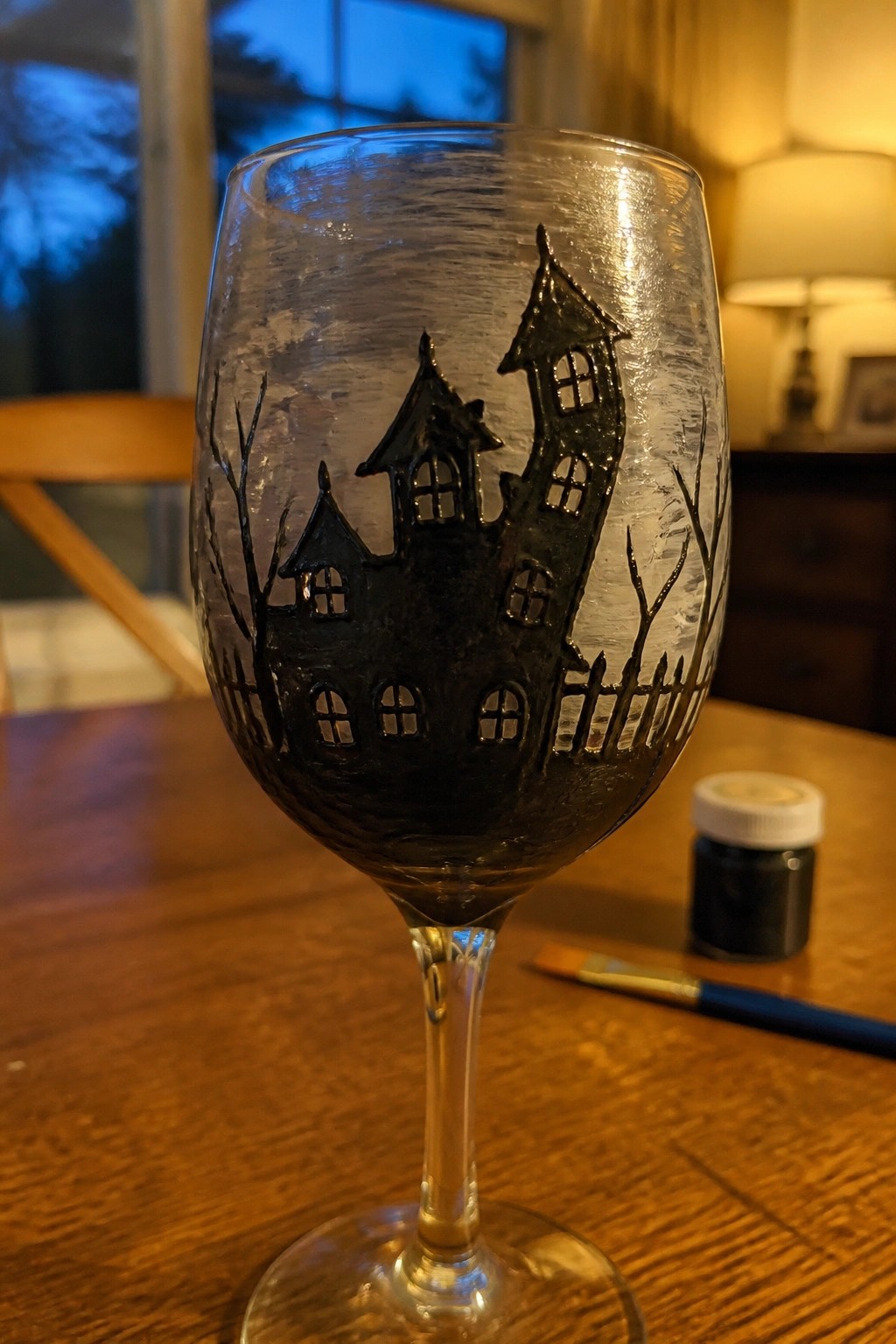Wine glass hand-painted with black haunted house, windows, trees, and fence.