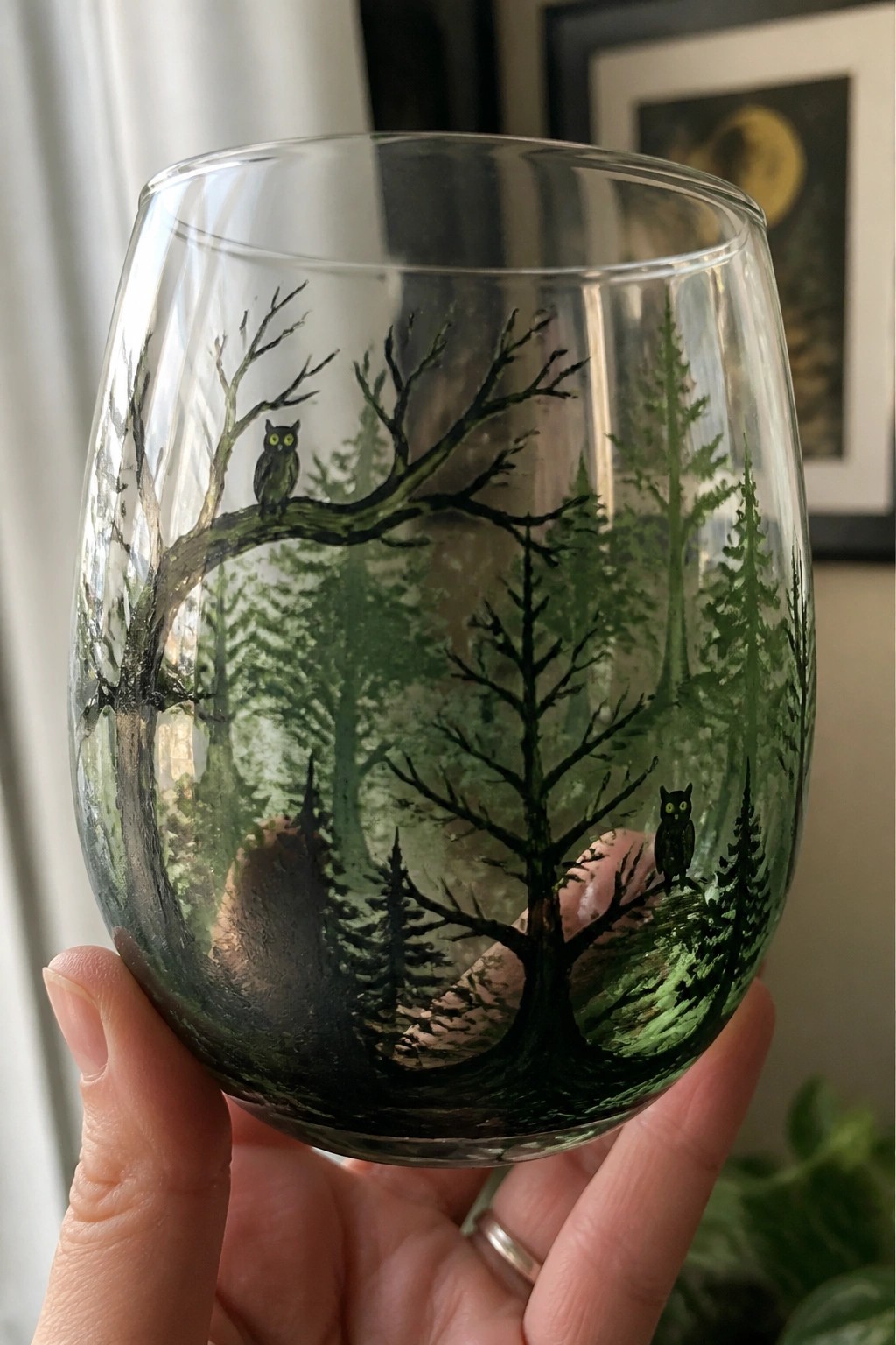 Hand holding clear glass painted with green forest, bare trees, and two owls.