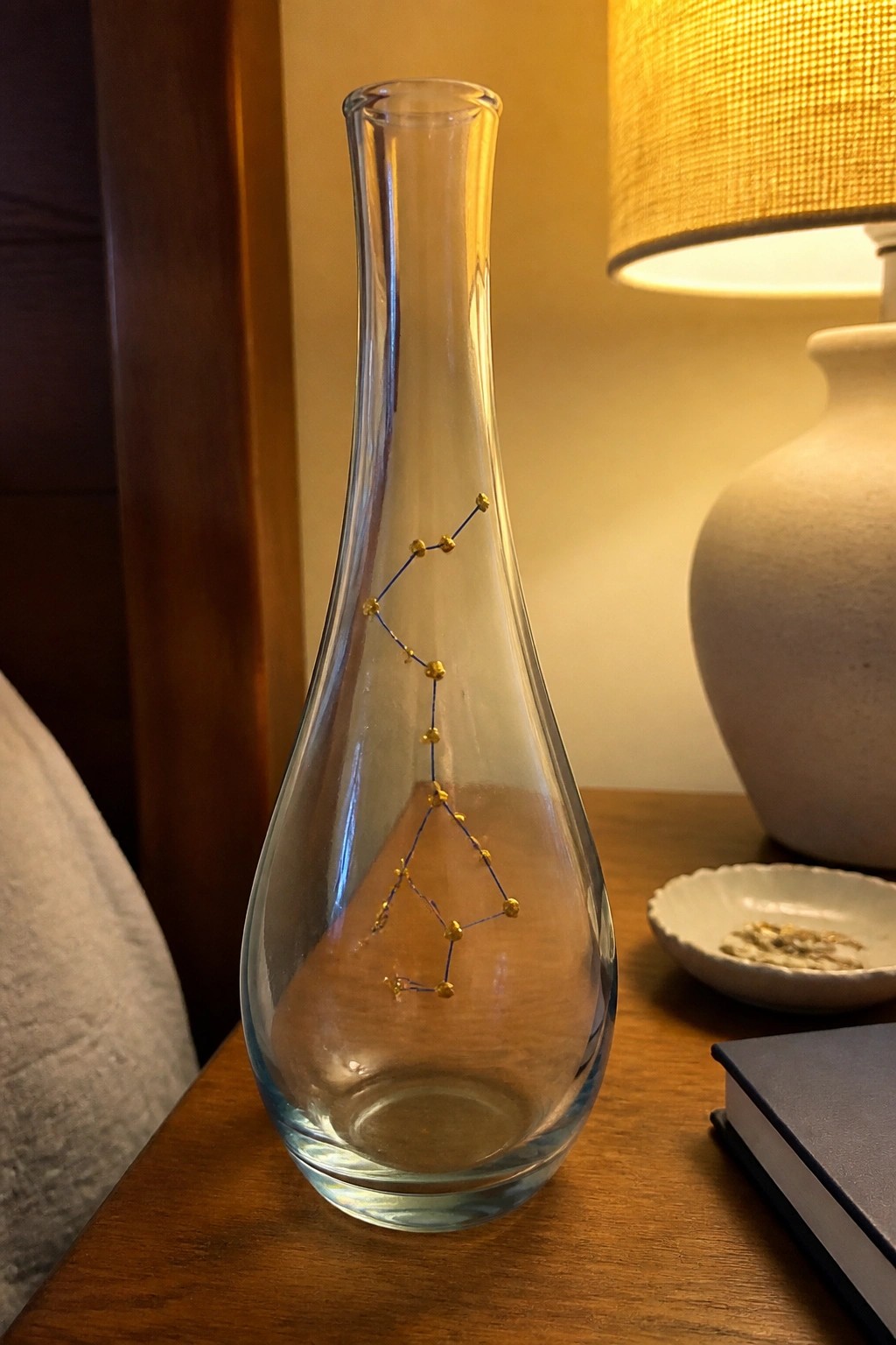 Tall glass vase with golden beaded constellation beside ceramic lamp on wood table